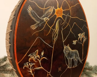 May include: A circular, hand-painted drum featuring a dark brown surface and an orange rim. The artwork showcases an eagle, a woman, a wolf, and lilies set against mountains and a sun. The drum's edges are bound with natural-coloured rope.