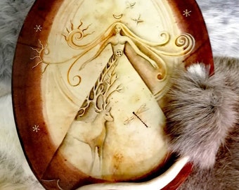 May include: A circular, brown leather wall hanging with a white center featuring a female figure with long flowing hair, a deer, a dragonfly, and various symbols. The background is a light brown color with a darker brown border.