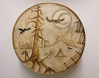 May include: A circular, brown, wooden drum with a painted design. The design features a tall, bare tree, a crescent moon, a flying owl, a tent, and several crows. The background is a light brown with a hazy, smoky effect.