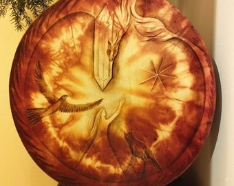 May include: A round, brown leather drum with a painted design. The design features a crystal, a bird, a woman, and a wolf. The background is a swirling pattern of light and dark brown.