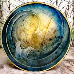 Könnte beinhalten: Ein runder Holzteller mit einem blauen und goldenen bemalten Design. Das Design zeigt zwei weiße Wölfe, die zu einem gelben Mond und Bergen im Hintergrund heulen.