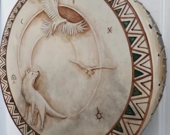May include: A white leather drum with a green and brown geometric pattern around the edge. The drum features a carved design of a wolf, a bird, and a sun symbol.