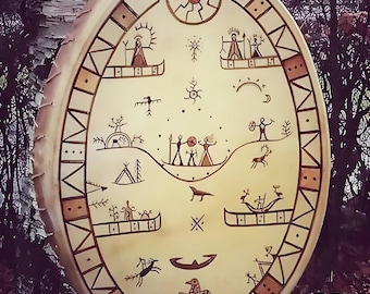 May include: A round, white leather drum with a brown painted design. The design features a variety of symbols, including a sun, a moon, stars, and animals. The drum is surrounded by a brown border with a geometric pattern.