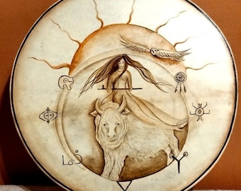May include: Round, hand-painted drum featuring a central image of a woman riding a white bison, with an owl and sun design. The drum has a light tan background with brown accents and symbols around the edge.