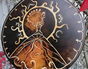 May include: A round wooden disc with a painted design of a stylized tree with a figure standing on a mountain. The tree has branches that extend to the edges of the disc. The figure is holding up its arms. The design is painted in gold and white on a black background.