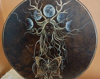 May include: A circular wooden panel with a dark brown background and a painted image of a woman with long flowing hair, a butterfly, and crescent moons. The woman is wearing a crown of antlers and the moons are in various phases.