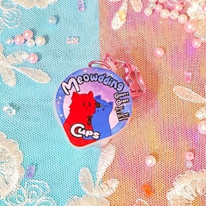 May include: A white charm with a purple background and two cartoon cats. The charm says "Meowdding" and "Galaxy Grape Cups".