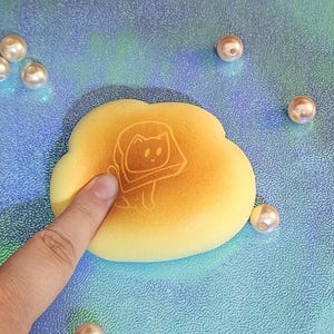 May include: A yellow squishy toy shaped like a cloud with a brown cat face inside. The cat face has a white outline and is looking to the right. The toy is on a blue and green iridescent surface.