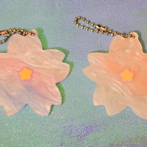 Op de afbeelding: Twee iriserende pastelroze en blauwe bloemenvormige sleutelhangers met een klein geel bloemetje in het midden. Elke sleutelhanger heeft een zilveren ketting.