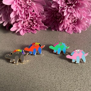 May include: Four enamel pins featuring a cartoon dinosaur with a rainbow colored back. The dinosaurs are red, green, gray, and pink.