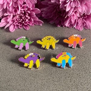 Puede incluir: Cinco pines de esmalte con forma de dinosaurio con rayas de diferentes colores en la espalda. Los colores de las rayas representan las diferentes banderas del orgullo.