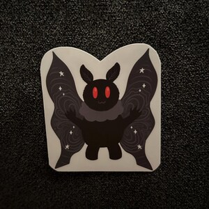 May include: A sticker featuring a cartoon moth with a black body, red eyes, and dark wings with white star accents. The moth has a gray collar and is standing on a white background.