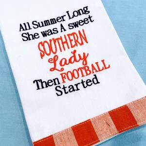 May include: White kitchen towel with an orange and white checkered border. The towel has an embroidered saying: "All Summer Long She Was A Sweet Southern Lady Then Football Started."