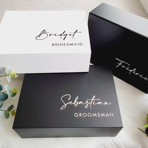 May include: Three square gift boxes, one white and two black, with names and titles printed on the lids. The white box reads "Bridget, Bridesmaid." The black boxes read "Frédéric" and "Sebastian, Groomsman."