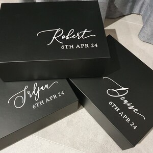 May include: Three black gift boxes with white lettering. The boxes are stacked on top of each other. The top box says "Robert 6TH APR 24". The middle box says "Seljan 6TH APR 24". The bottom box says "Denise 6TH APR 24".