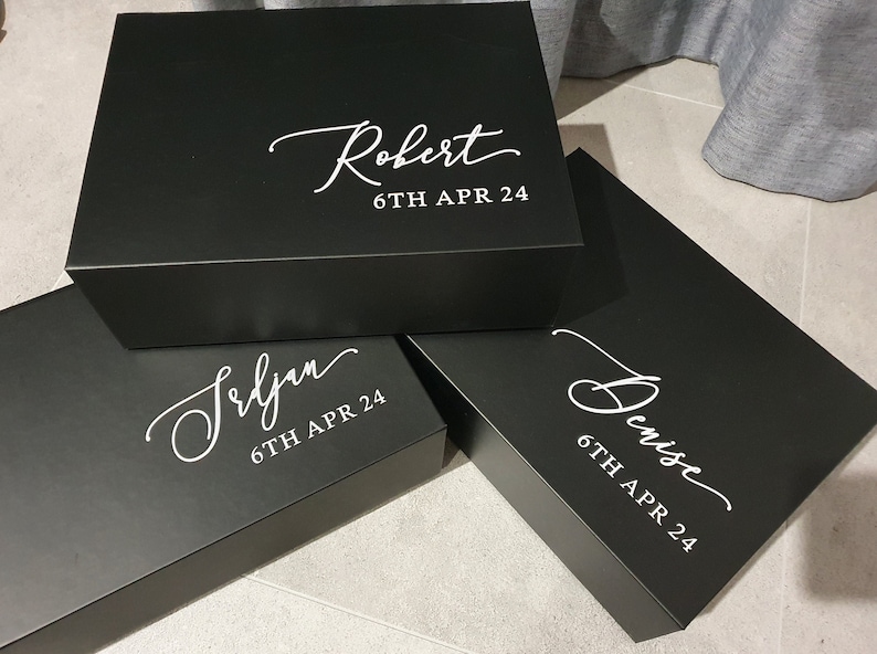 May include: Three black gift boxes with white lettering. The boxes are stacked on top of each other. The top box says "Robert 6TH APR 24". The middle box says "Seljan 6TH APR 24". The bottom box says "Denise 6TH APR 24".