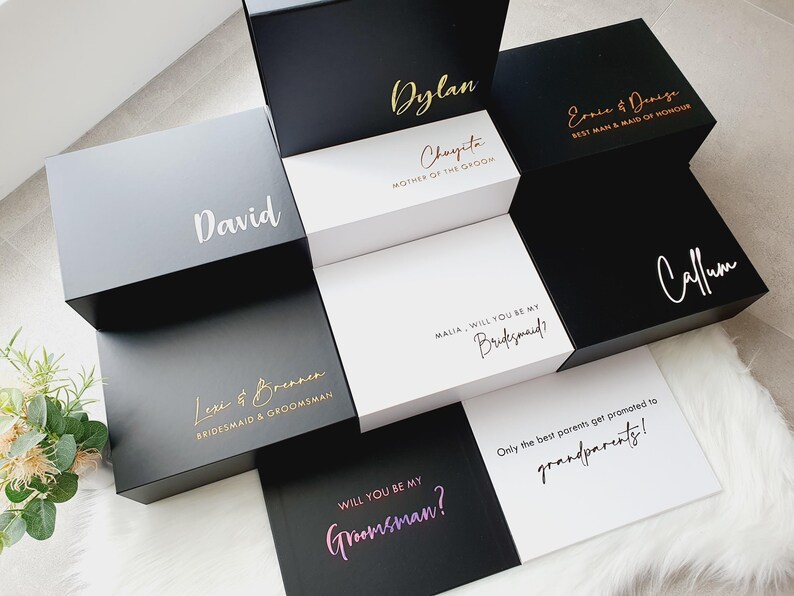 K&ouml;nnte beinhalten: Ein Set aus schwarzen und wei&szlig;en Geschenkboxen mit goldenen Schriftz&uuml;gen. Die Boxen sind mit Namen und Titeln wie "Best Man", "Maid of Honor", "Mother of the Groom" und "Groomsman" verziert.