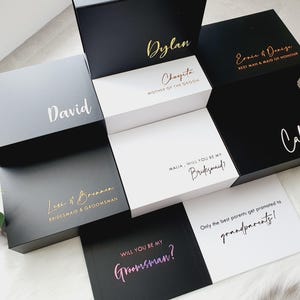May include: A set of black and white gift boxes with gold lettering. The boxes are decorated with names and titles such as "Best Man", "Maid of Honour", "Mother of the Groom", and "Groomsman".
