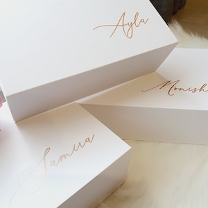 May include: Three white gift boxes stacked on top of each other. The top box has the name "Ayla" written in gold script. The middle box has the name "Monisha" written in gold script. The bottom box has the name "Samira" written in gold script.