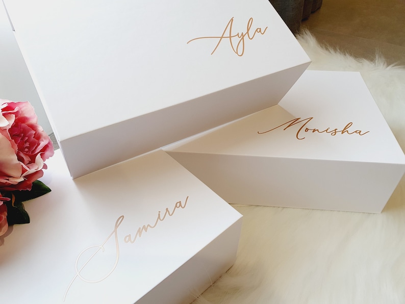 May include: Three white gift boxes stacked on top of each other. The top box has the name "Ayla" written in gold script. The middle box has the name "Monisha" written in gold script. The bottom box has the name "Samira" written in gold script.