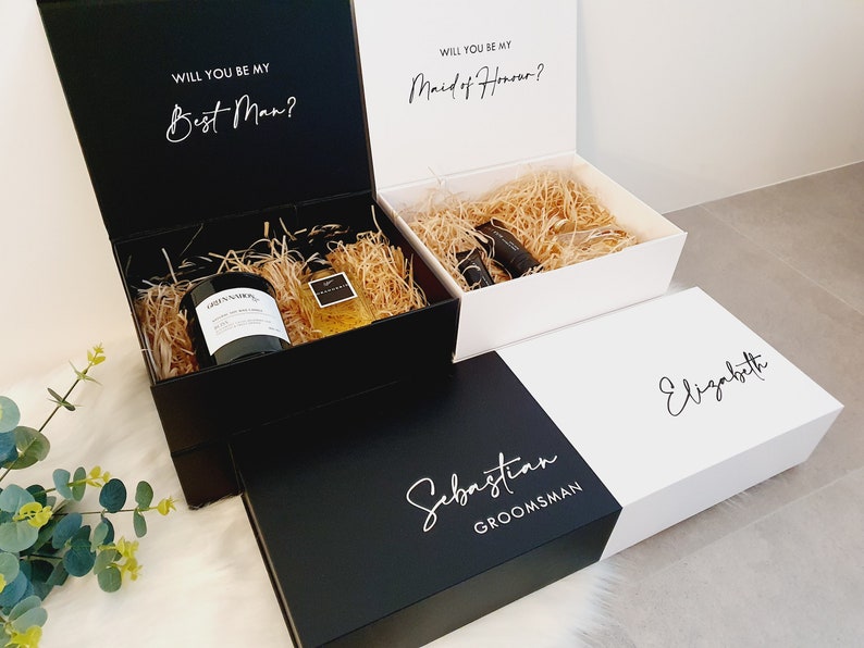 K&ouml;nnte beinhalten: Zwei Geschenkboxen mit ge&ouml;ffneten Deckeln, eine schwarze und eine wei&szlig;e. Die schwarze Box enth&auml;lt eine Kerze und eine Flasche Fl&uuml;ssigkeit. Die wei&szlig;e Box enth&auml;lt eine Flasche Fl&uuml;ssigkeit. Die Boxen haben den Text "Will you be my Best Man?" und "Will you be my Maid of Honour?" auf den Deckeln. Die Boxen haben auch die Namen "Sebastian" und "Elizabeth" auf der Vorderseite.