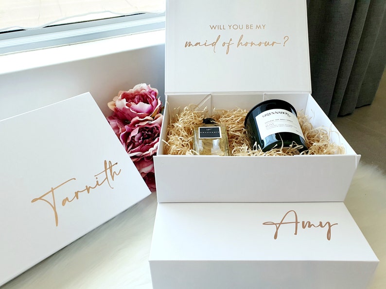 May include: White gift boxes with gold lettering. The top box says "Will you be my maid of honour?" The middle box says "Tarnitih" and the bottom box says "Amy". The boxes contain a candle and a bottle of perfume.