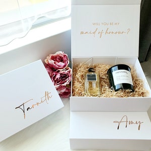 K&ouml;nnte beinhalten: Zwei wei&szlig;e Geschenkboxen mit goldener Beschriftung. Die obere Box tr&auml;gt den Text "Willst du meine Trauzeugin sein?" Die untere Box tr&auml;gt den Namen "Amy". Die mittlere Box ist ge&ouml;ffnet und enth&auml;lt ein Parf&uuml;mfl&auml;schchen und eine schwarze Kerze.