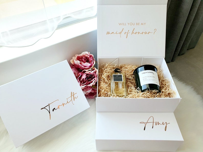 K&ouml;nnte beinhalten: Zwei wei&szlig;e Geschenkboxen mit goldener Beschriftung. Die obere Box tr&auml;gt den Text "Willst du meine Trauzeugin sein?" Die untere Box tr&auml;gt den Namen "Amy". Die mittlere Box ist ge&ouml;ffnet und enth&auml;lt ein Parf&uuml;mfl&auml;schchen und eine schwarze Kerze.