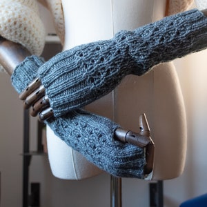 Puede incluir: Un par de guantes sin dedos grises con un patrón de punto texturizado. Los guantes se muestran en un maniquí de mano de madera.
