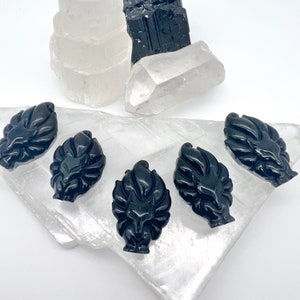 May include: Five black obsidian fox carvings are arranged on a white selenite crystal slab, with additional selenite, quartz, and black tourmaline crystals in the background. The fox carvings are detailed and polished.