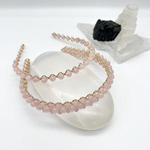 May include: Two gold wire headbands with pink rose quartz beads. The headbands are on a white surface.