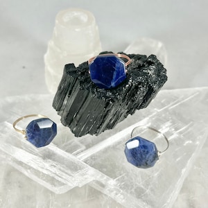 May include: Three rings with blue gemstone settings. The rings are made of gold and silver wire and feature different sized hexagonal shaped stones. The rings are displayed on a white crystal surface.