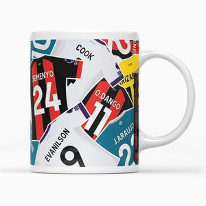 Può includere: Una tazza in ceramica bianca con un design colorato di maglie da calcio con numeri e nomi. Le maglie sono di squadre diverse e presentano vari colori, tra cui rosso, nero, bianco, blu e giallo.