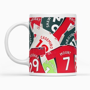 May include: White ceramic mug featuring a design of red and green football shirts with player names and numbers. The shirts are layered on top of each other, creating a collage effect. The names on the shirts include "Amrabat", "Shaw", "Casemiro", "Hojlund", "Mount", "Bissaka", and "Bayindir".