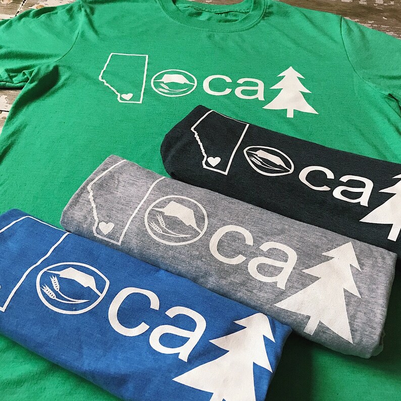 K&ouml;nnte beinhalten: Vier gefaltete T-Shirts mit einem wei&szlig;en grafischen Design aus einem Kiefernbaum, einem Herzen, einem Weizenhalm und einer staatlichen Umrandung. Der Text "loca ca" ist auf jedem T-Shirt aufgedruckt. Die T-Shirts sind in verschiedenen Farben: gr&uuml;n, schwarz, grau und blau.