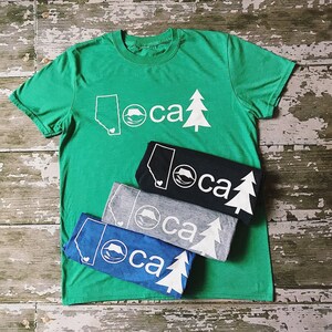 K&ouml;nnte beinhalten: Vier T-Shirts &uuml;bereinander gestapelt. Das oberste Shirt ist gr&uuml;n mit einem wei&szlig;en Umriss einer Bergkette, eines B&auml;ren und einer Kiefer. Der Text "loca" ist in Wei&szlig; gedruckt. Die anderen Shirts sind schwarz, grau und blau mit dem gleichen Design in Wei&szlig;.
