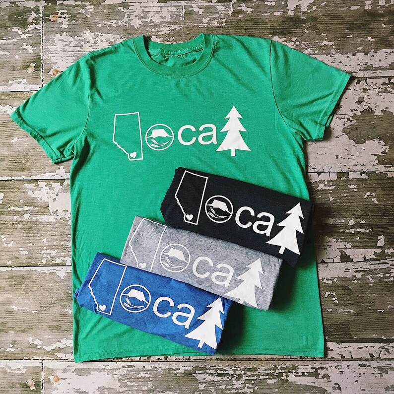 K&ouml;nnte beinhalten: Vier T-Shirts &uuml;bereinander gestapelt. Das oberste Shirt ist gr&uuml;n mit einem wei&szlig;en Umriss einer Bergkette, eines B&auml;ren und einer Kiefer. Der Text "loca" ist in Wei&szlig; gedruckt. Die anderen Shirts sind schwarz, grau und blau mit dem gleichen Design in Wei&szlig;.