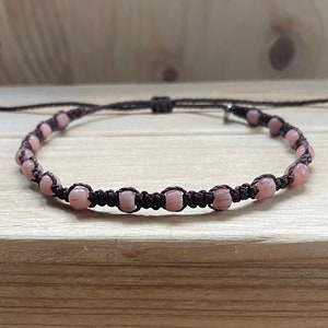 Könnte beinhalten: Ein braunes Kordel-Armband mit rosa Perlen. Das Armband ist mit einem Knoten gebunden und hat einen silbernen Verschluss.