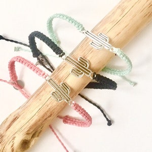 Pode incluir: Quatro pulseiras de amizade com um amuleto de cacto. As pulseiras são feitas de fio rosa, preto e verde. O amuleto de cacto é prateado.