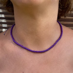 Puede incluir: Un collar de cuentas moradas con un diseño sencillo. El collar está hecho de pequeñas cuentas redondas que están enhebradas en un cordón delgado.