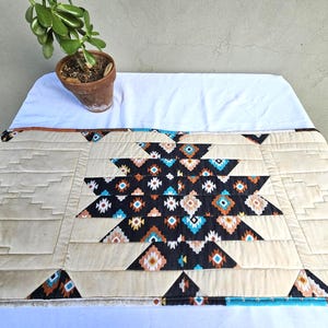 May include: A beige and black geometric patterned table runner with a central design of triangles and diamonds in shades of black, turquoise, and orange. The runner is quilted and has a border of brown fabric.