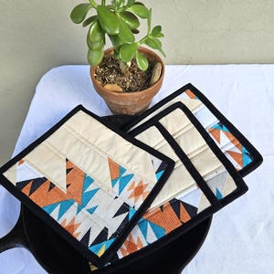 May include: Four square fabric coasters with a geometric design in teal, orange, black, and white, are placed in a black cast iron skillet. A small potted jade plant sits nearby. The coasters have a cream-colored top with a black border.