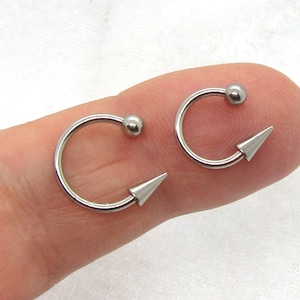 May include: Two silver-toned nose rings with a pointed end and a small ball on the other end. The rings are shown on a finger.