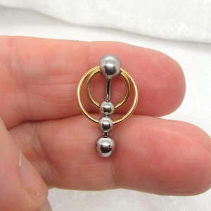 May include: A silver and gold-toned body piercing jewelry piece. The jewelry features a barbell design with three silver balls and two gold-toned rings. The jewelry is held in a person's hand.