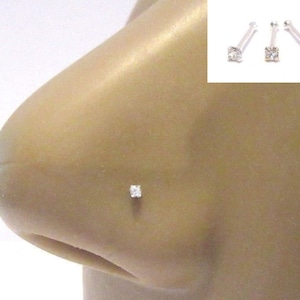 May include: A close-up of a nose piercing with a small, clear, square-shaped gemstone. The piercing is made of silver and is a popular choice for nose jewellery.