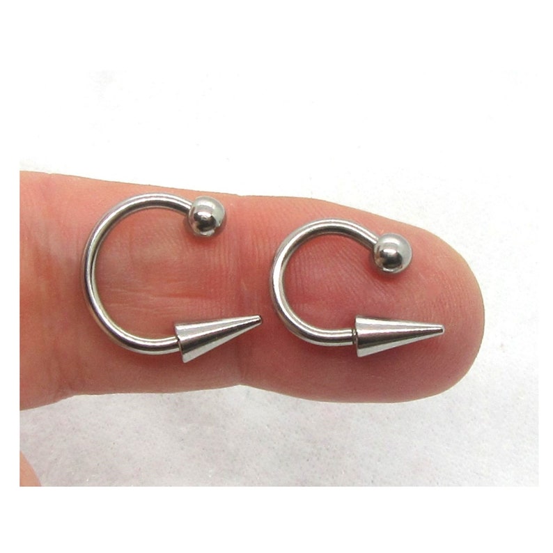 Puede incluir: Dos piezas de joyer&iacute;a corporal plateadas con un aro circular y un extremo puntiagudo. El aro tiene una peque&ntilde;a bola en la parte superior.