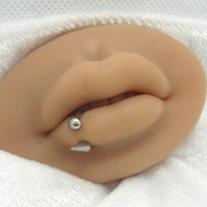 Puede incluir: Primer plano de la boca de una persona con un aro de labio plateado. El aro tiene una peque&ntilde;a bola en un extremo y un extremo puntiagudo en el otro.