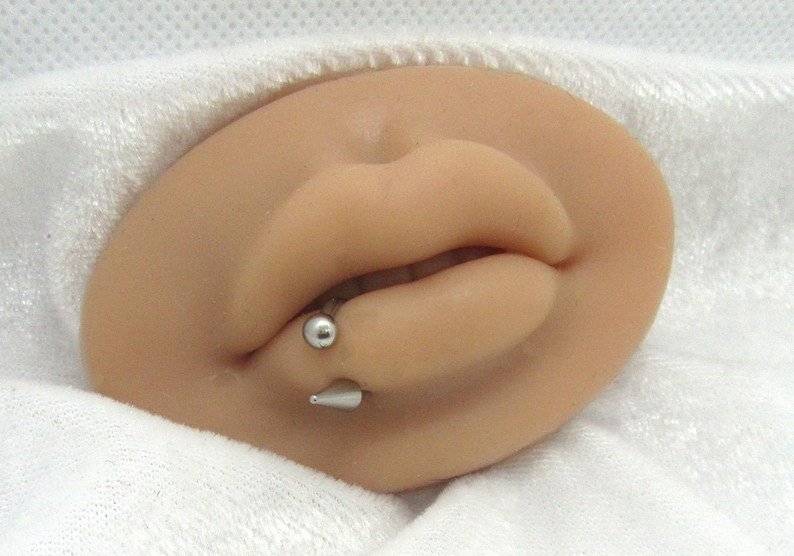 Puede incluir: Primer plano de la boca de una persona con un aro de labio plateado. El aro tiene una peque&ntilde;a bola en un extremo y un extremo puntiagudo en el otro.