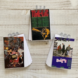 May include: Three small spiral-bound notebooks with album art covers. One features "TONE-LOC" in red and green text. Another has "BELL BIV DEVOE" and "POISON" text. The third notebook displays "BUCKS ZDAGIC" text and a colorful illustration.