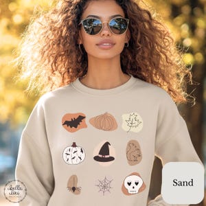 May include: A sand-coloured crewneck sweatshirt featuring a grid of nine autumn and Halloween-themed line art illustrations, including a bat, pumpkin, maple leaf, witch hat, squirrel, spider, spiderweb, and a skull, set against soft-toned shapes.